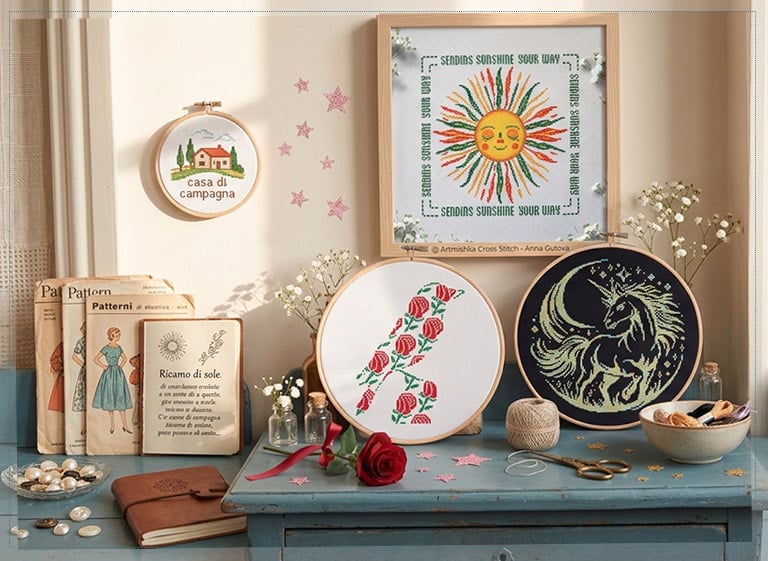 Artmishka cross stitch pieces on a vintage table: smiling sun, floral initial and unicorn, among pattern booklets and wildflowers.